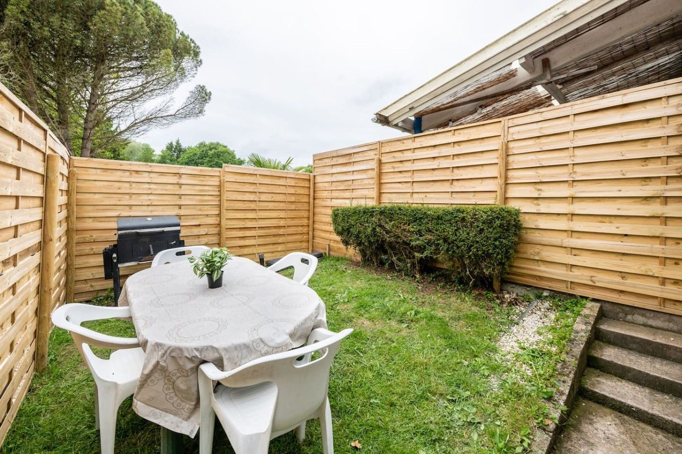 Un jardin avec une table ronde, des chaises blanches, un barbecue et une clôture en bois.