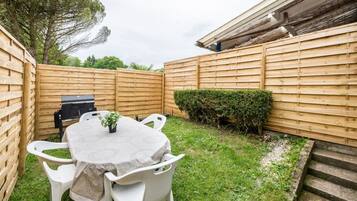 Un jardin avec une table ronde, des chaises blanches, un barbecue et une clôture en bois.