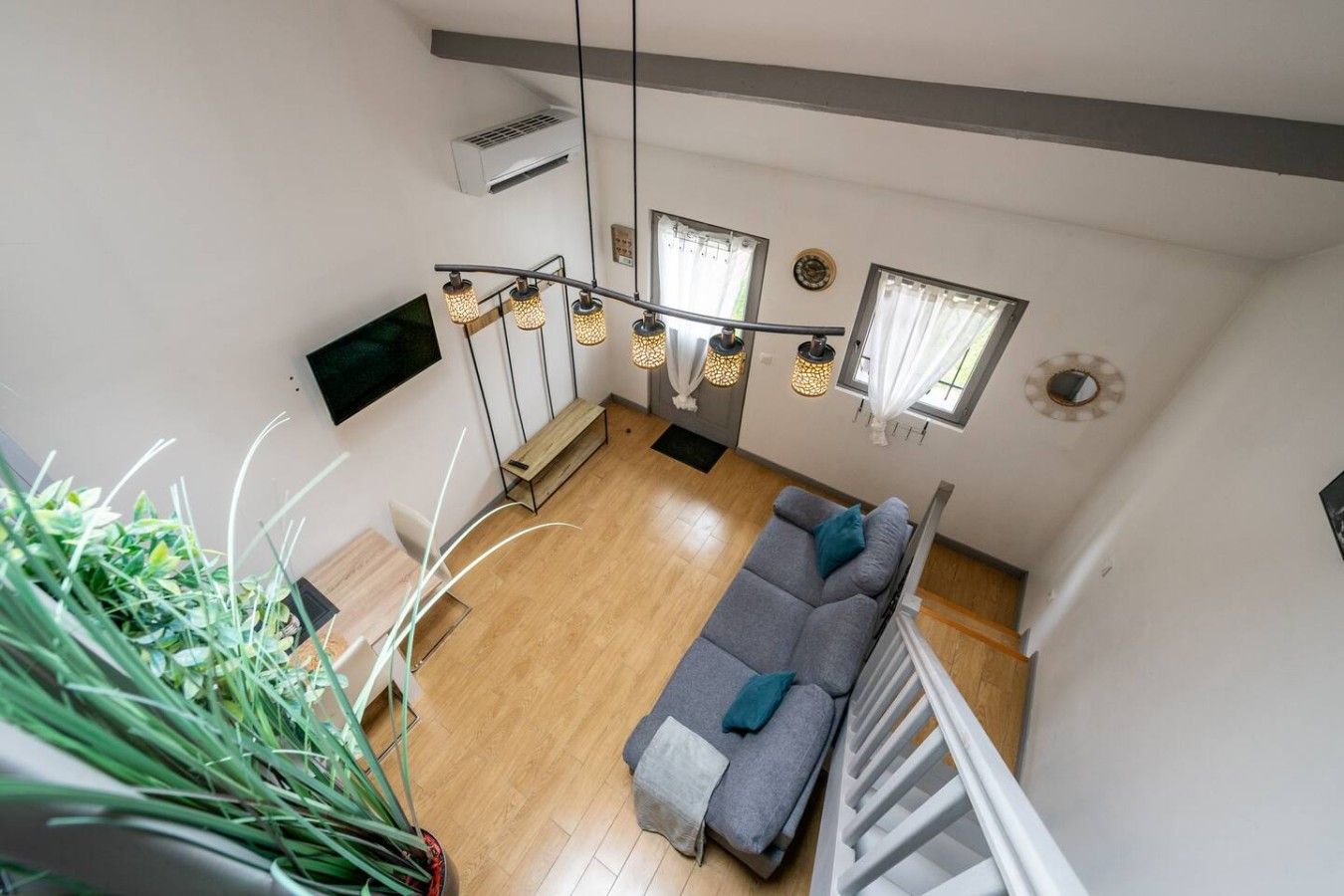 A high-ceiling living room with a wooden floor, a grey sofa, and a staircase with a white railing.