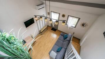 A high-ceiling living room with a wooden floor, a grey sofa, and a staircase with a white railing.