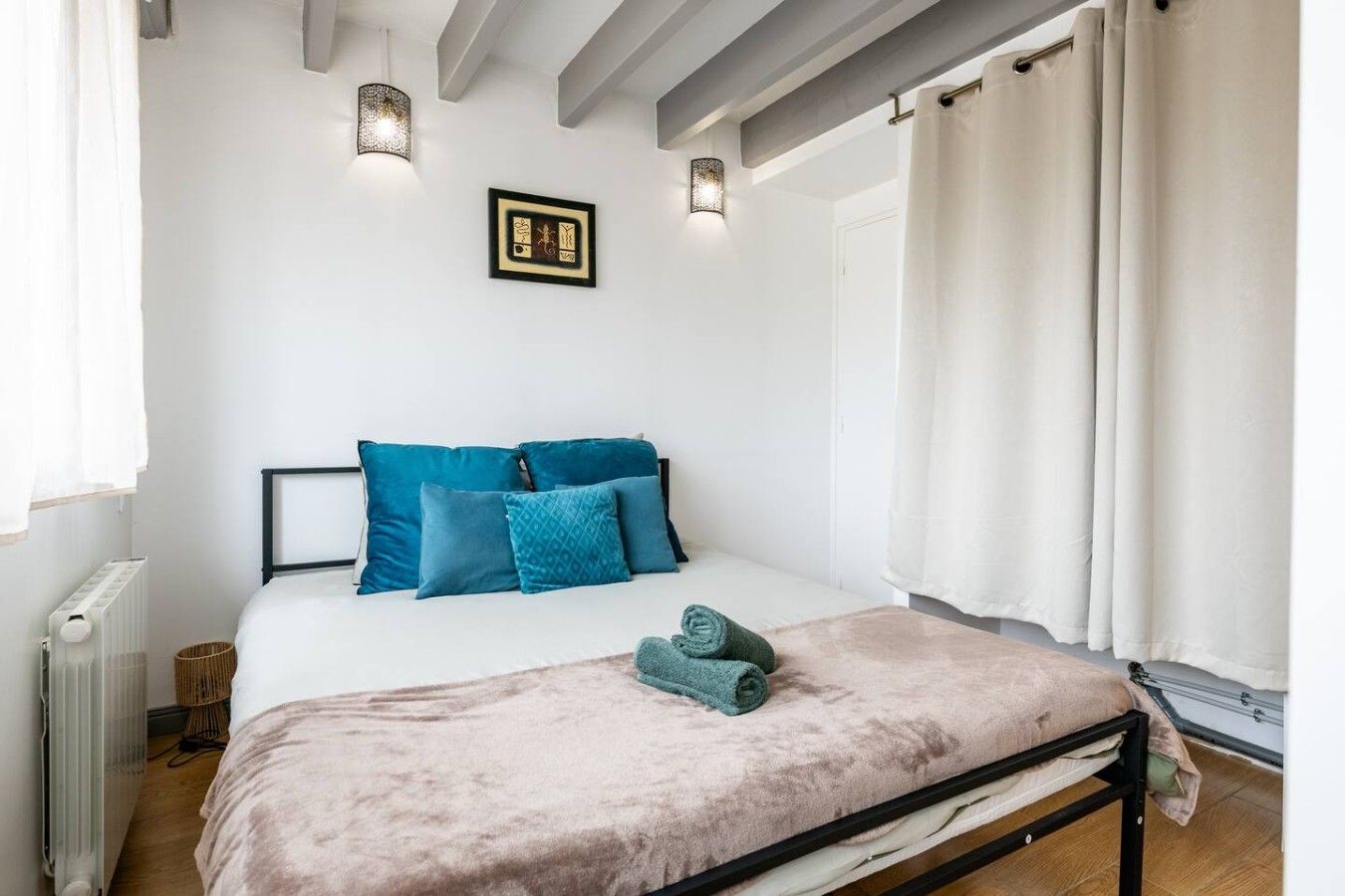 Un dormitorio con cama, almohadas, una colcha, un cuadro en la pared y una ventana con cortinas.