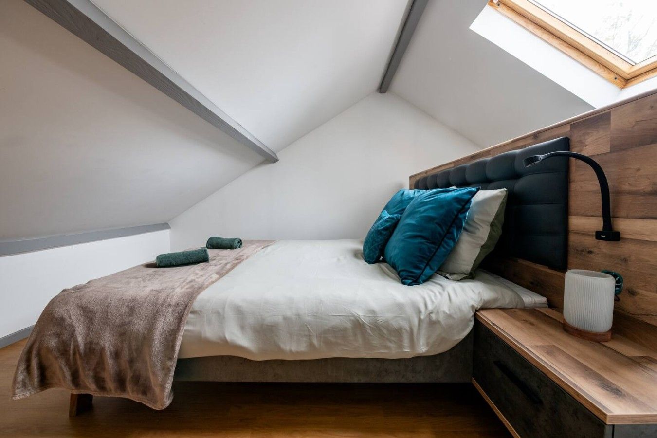 Een slaapkamer met een schuine kap, een bed met hoofdeinde, kussens, een nachtkastje met een lamp en een raam.