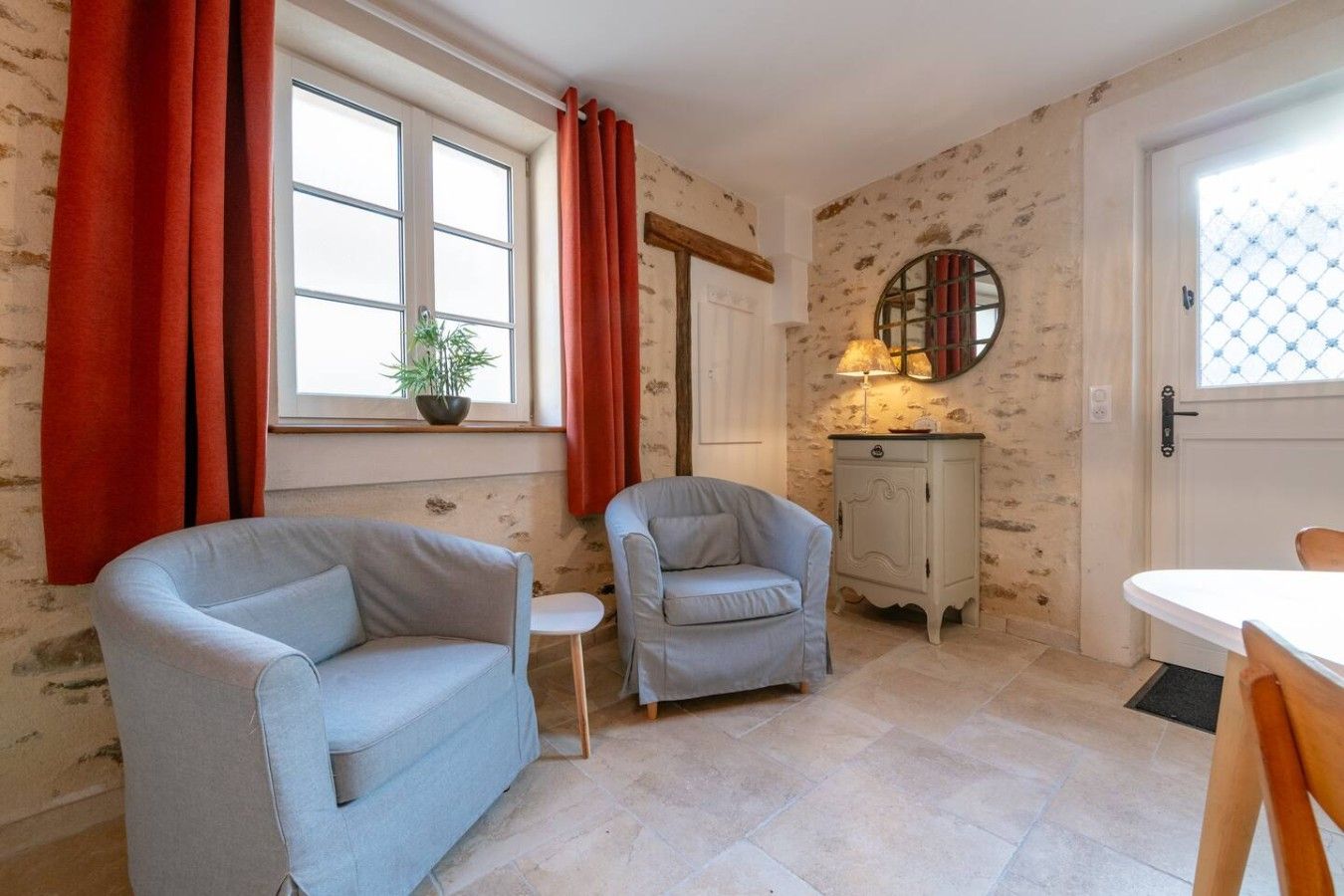 A room with two light-colored armchairs, a small white table, a cabinet, and a window with red curtains.