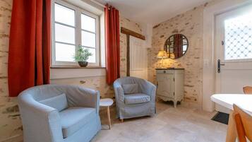 A room with two light-colored armchairs, a small white table, a cabinet, and a window with red curtains.