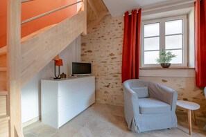 A room with a white dresser, a television on top, a red curtain, a window with a plant, and a light blue chair. - La Blandynette—renovated and charming (Blandy)