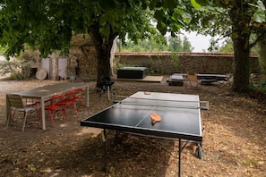 Tischtennis-Ausrüstung im Freien mit Tisch, Stühlen und Schläger, umgeben von Bäumen und einer rustikalen Steinmauer.