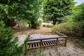 A wooden bench with a patterned cushion in a garden area with gravel ground, surrounded by dense green foliage. - House, Lot | Hot Tub • Fire pit • Art & Deco (Bourdenay)
