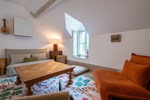 A bedroom with a bed, a wooden table, a sofa, a lamp, a window, and a guitar on the wall. - House, Lot | Hot Tub • Fire pit • Art & Deco (Bourdenay)