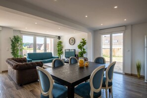 A modern living room with a sofa, a dining table with chairs, and a TV. There are large windows and a view of the outside. - Le Panoramique | Exceptional Home • 12 trips (Crécy-la-Chapelle)