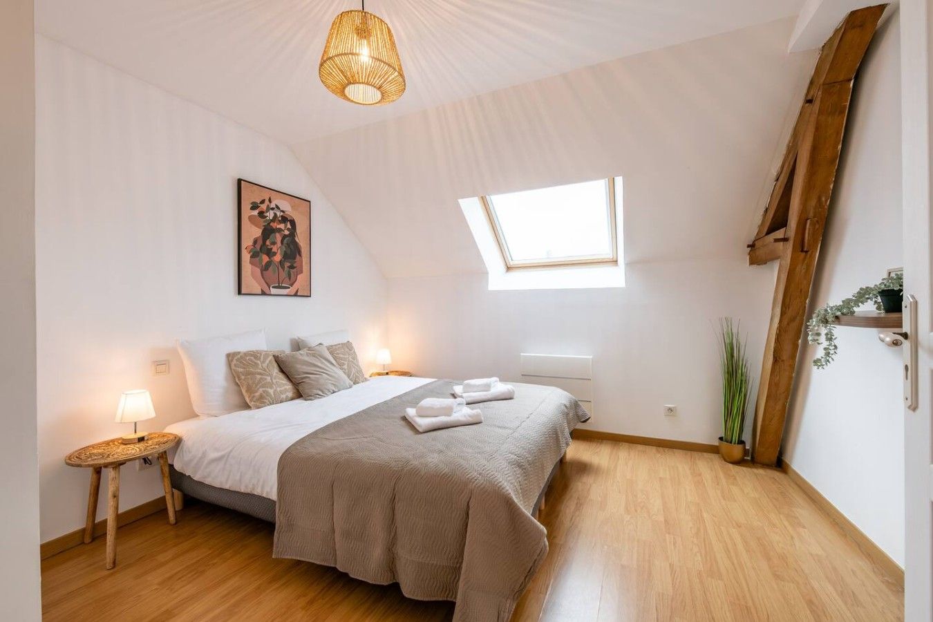 Un dormitorio con cama, lámparas de noche, tragaluz, escalera de madera y un cuadro en la pared.