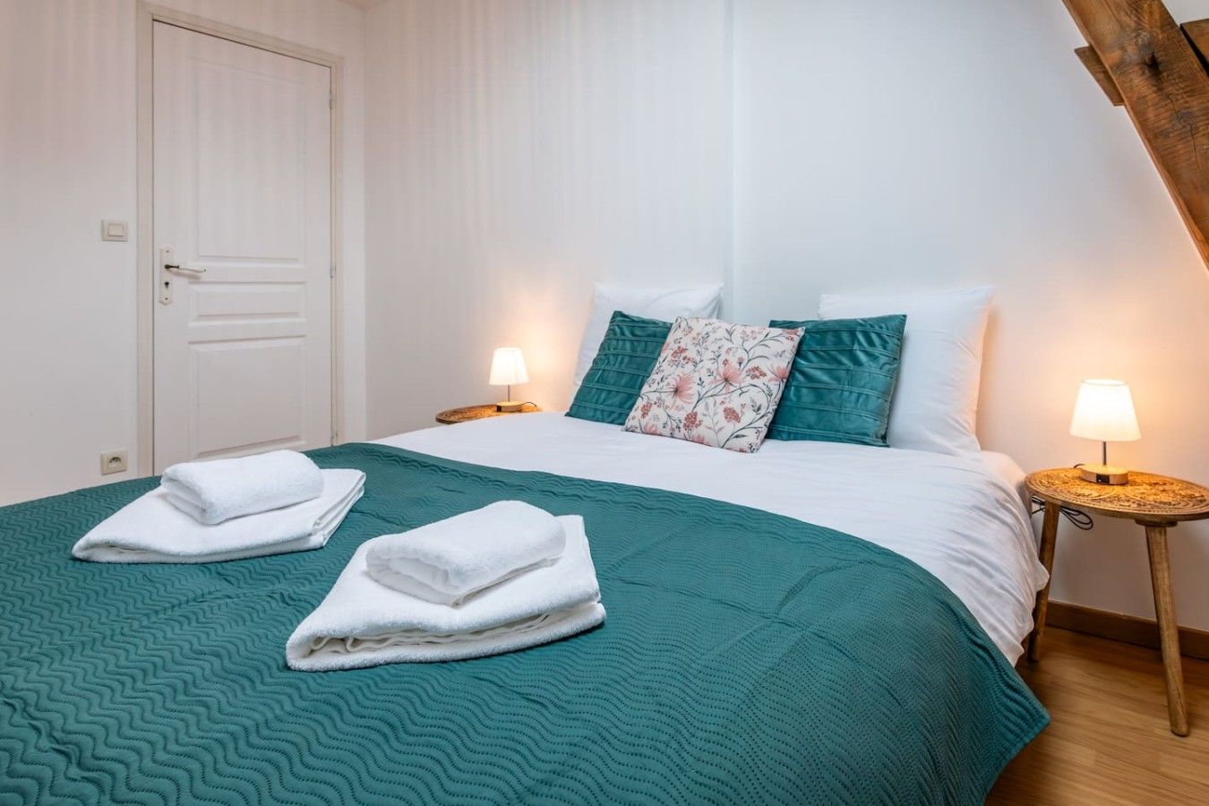 A neatly made bed with two pillows, a teal bedspread, and two folded towels. There are two bedside lamps on wooden nightstands.