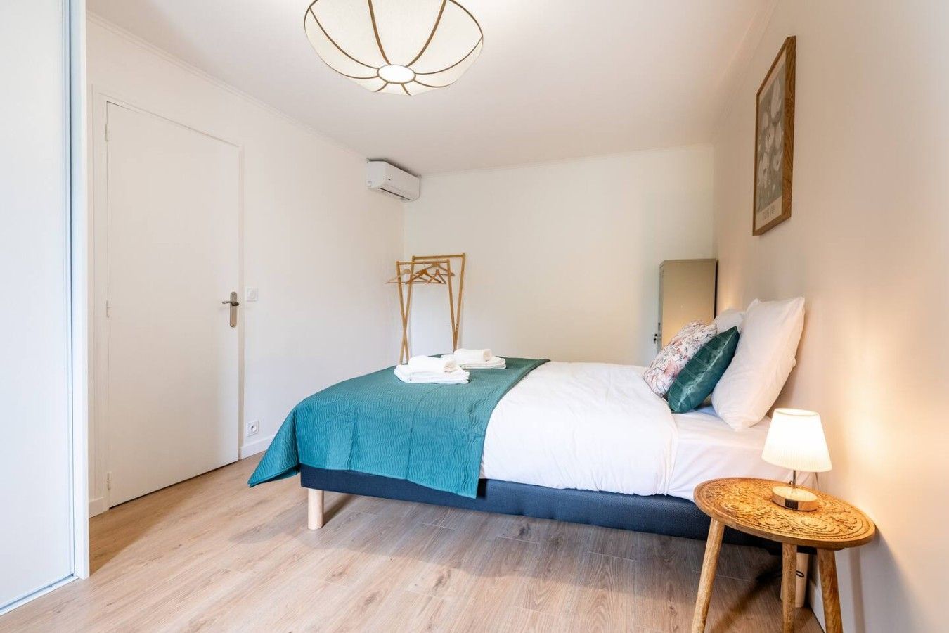 Ein Schlafzimmer mit einem Bett, einem Nachttisch mit Lampe, einem Kleiderständer und einem Bild an der Wand.