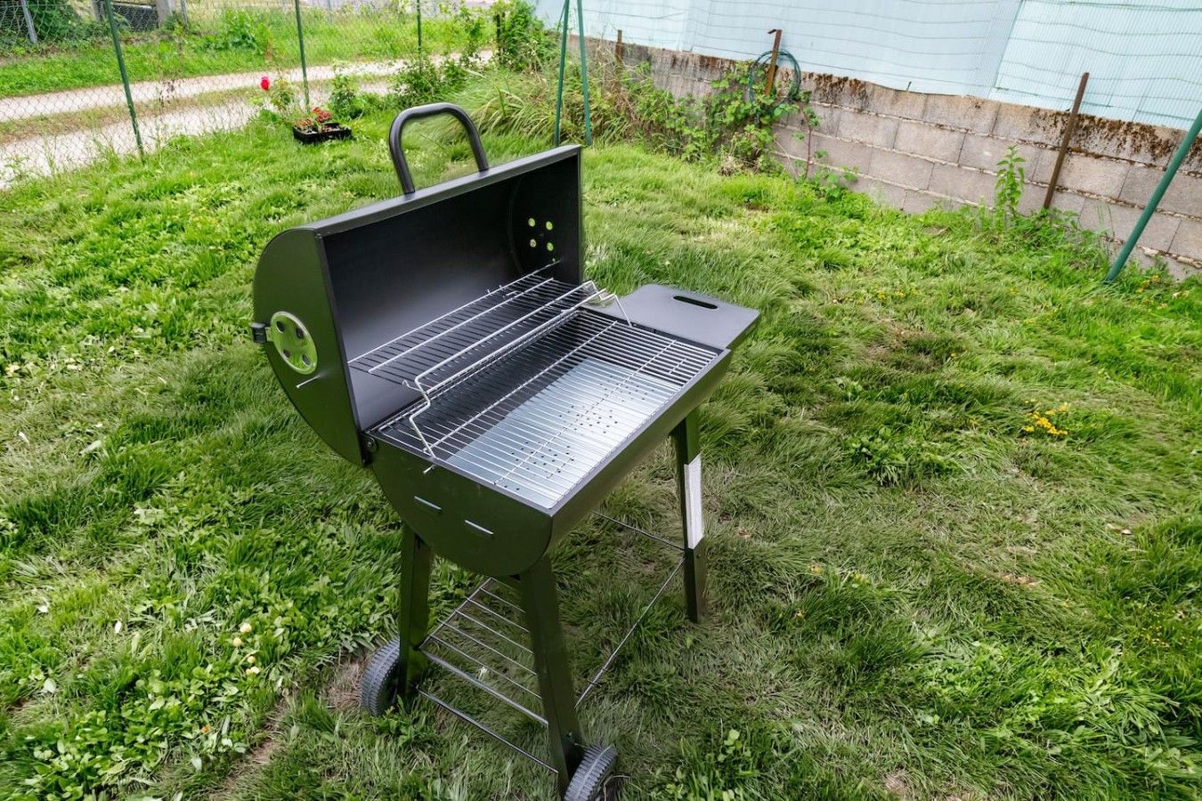 A portable grill with a wire rack, set on grass with a green fence and plants in the background.