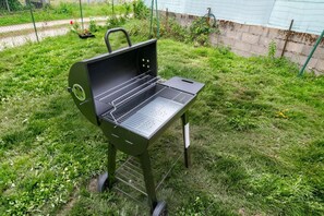 A portable grill with a wire rack, set on grass with a green fence and plants in the background. - A Little Getaway—Air-conditioned home, barbecue, renovated (Longueville)