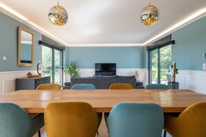 A modern living room with a large wooden dining table, two sofas, a TV, and large windows. - L'Évasion – House with pool and hot tub, sleeps 14 (Lescherolles)