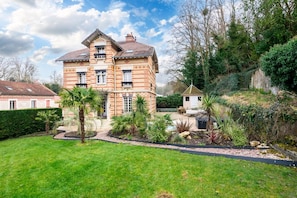A two-story house with a brick facade, a small attached garage, and a well-maintained garden with various plants and a lawn. - Villa Les Roches - Comfort - Relaxation - Pool (La Ferté-Gaucher)