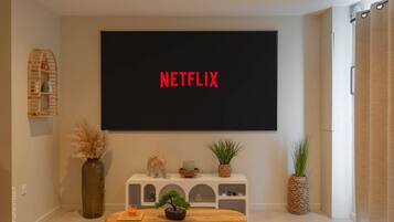 Un soggiorno con una grande TV a schermo piatto montata a parete che mostra 'NETFLIX' in rosso, un piccolo tavolo consolle bianco con oggetti decorativi, un tavolino in legno e una pianta in vaso.