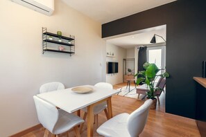Un comedor moderno con una mesa blanca, cuatro sillas blancas, una estantería negra con plantas y una planta verde grande en la esquina.
