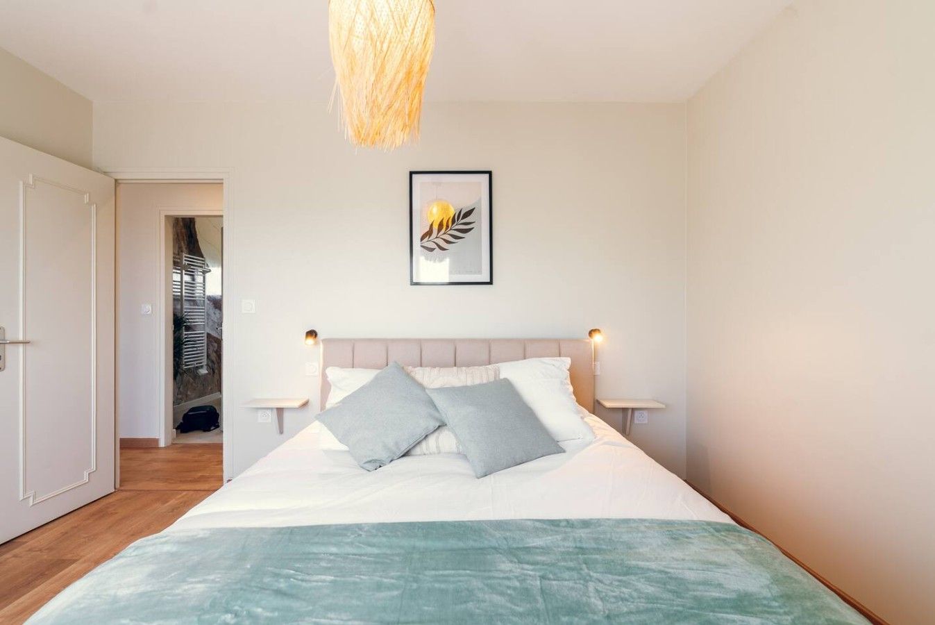 Une chambre à coucher avec un lit, des lampes de chevet, un tableau encadré et une porte donnant sur une autre pièce.