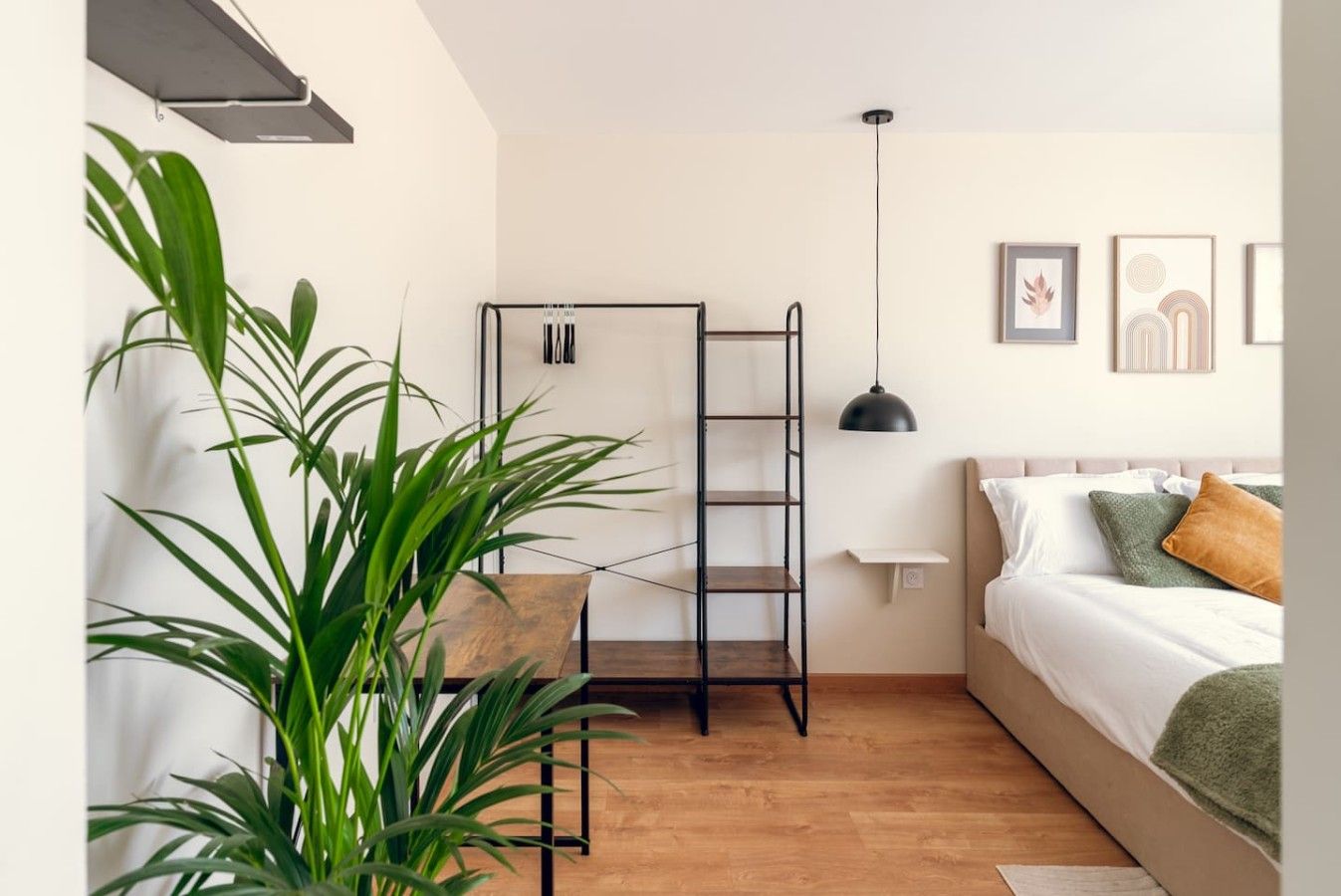 A bedroom with a bed, a wooden floor, a black metal shelf, a hanging pendant light, and a potted plant.