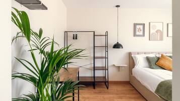 A bedroom with a bed, a wooden floor, a black metal shelf, a hanging pendant light, and a potted plant.