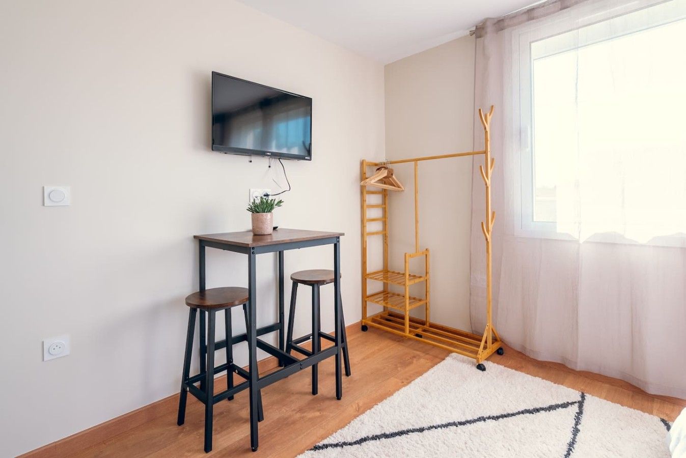 A room with a TV mounted on the wall, a small table with stools, a coat rack, and a window with curtains.