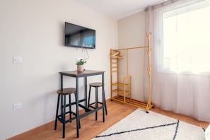 A room with a TV mounted on the wall, a small table with stools, a coat rack, and a window with curtains.