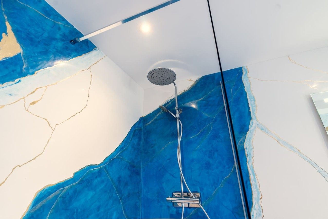 Een moderne badkamer met een grote blauwe marmeren wand, een in het plafond gemonteerde douchekop en zichtbare leidingen.