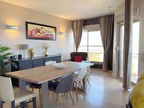 A modern dining area with a wooden table, white chairs, a black sofa, and a large window with curtains. - Stunning Beachfront 3 Bedroom Apartment in Valras Plage with Balcony (Valras-Plage)