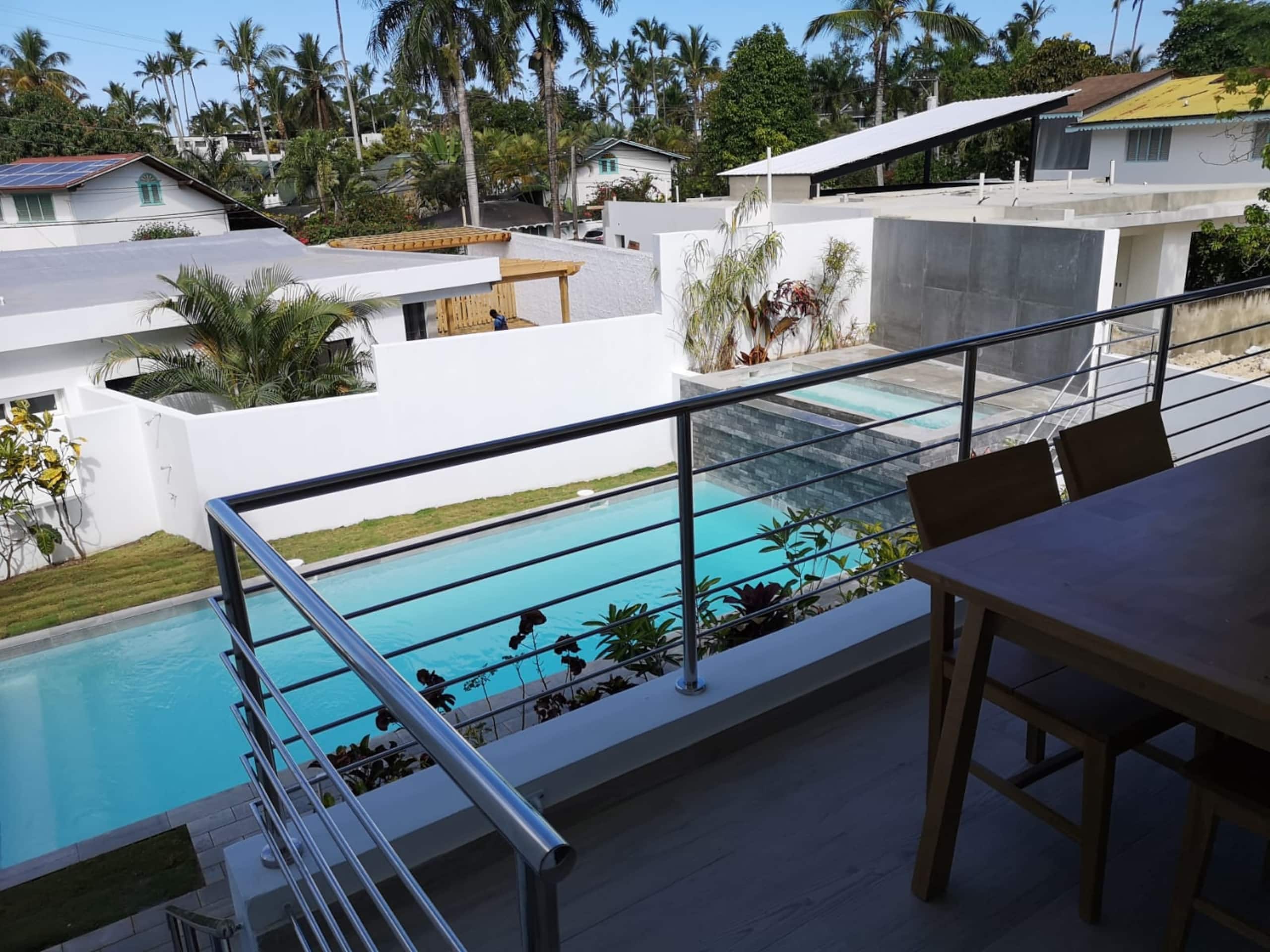 A modern residential area with a swimming pool, a balcony with a table and chairs, and a view of neighboring houses and palm trees.