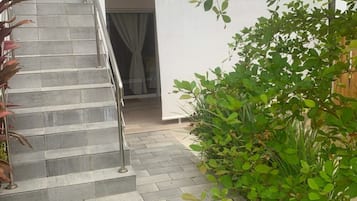 Escadaria que leva a um prédio branco com guarda-corpo de metal, cercado por vegetação e um chão de azulejos.
