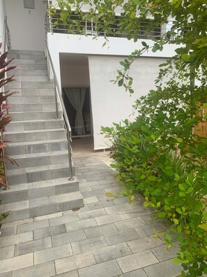 Een trap leidt naar een wit gebouw met een metalen leuning, omgeven door groen en een betegelde vloer.