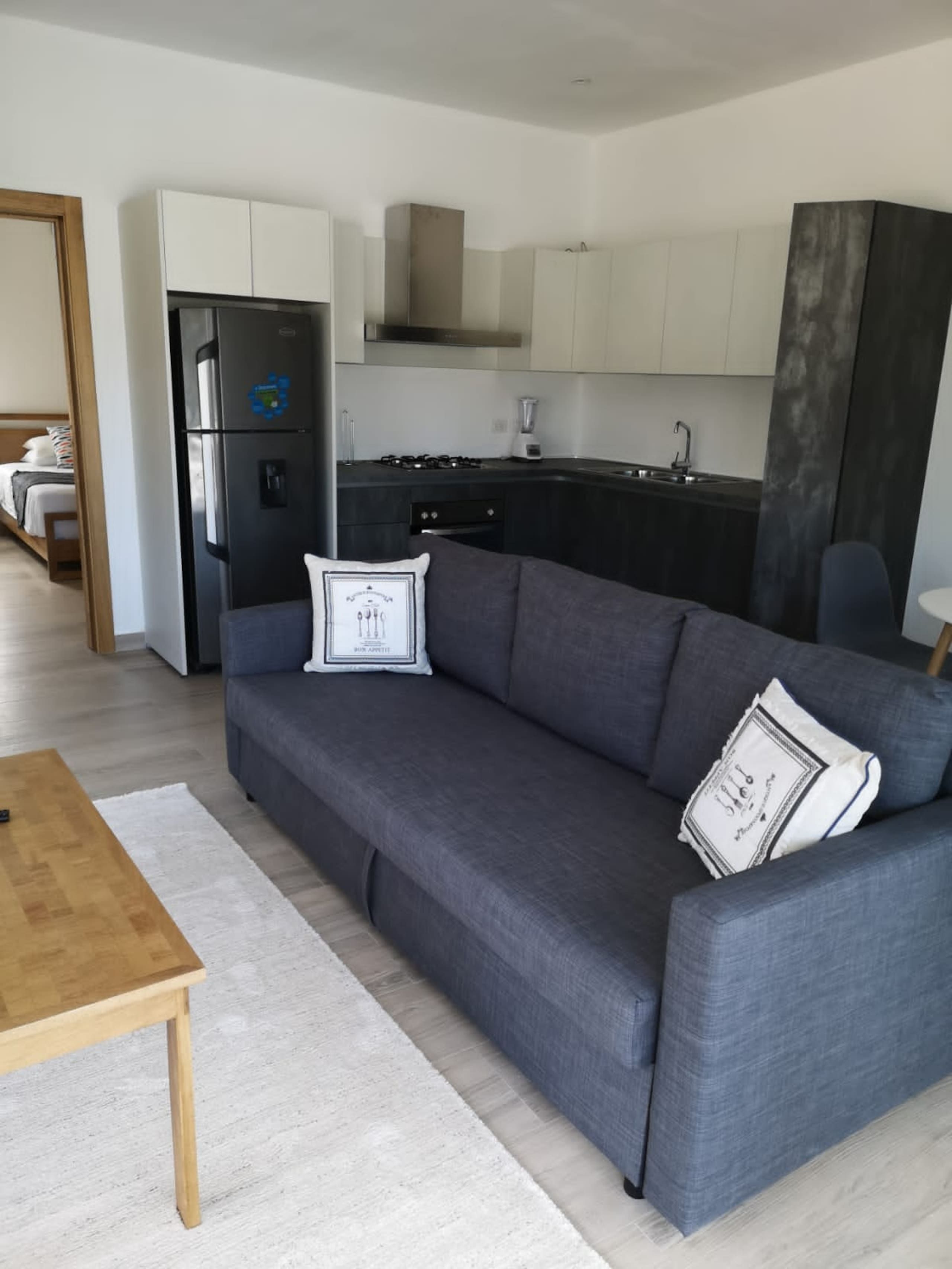 Una sala moderna con un sofá gris, una mesa de centro de madera y una zona de cocina con armarios blancos y un refrigerador negro.