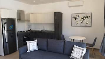 Cozinha moderna com fogão embutido, geladeira e pia. Há um sofá cinza com almofadas decorativas e uma pequena mesa redonda com cadeiras.