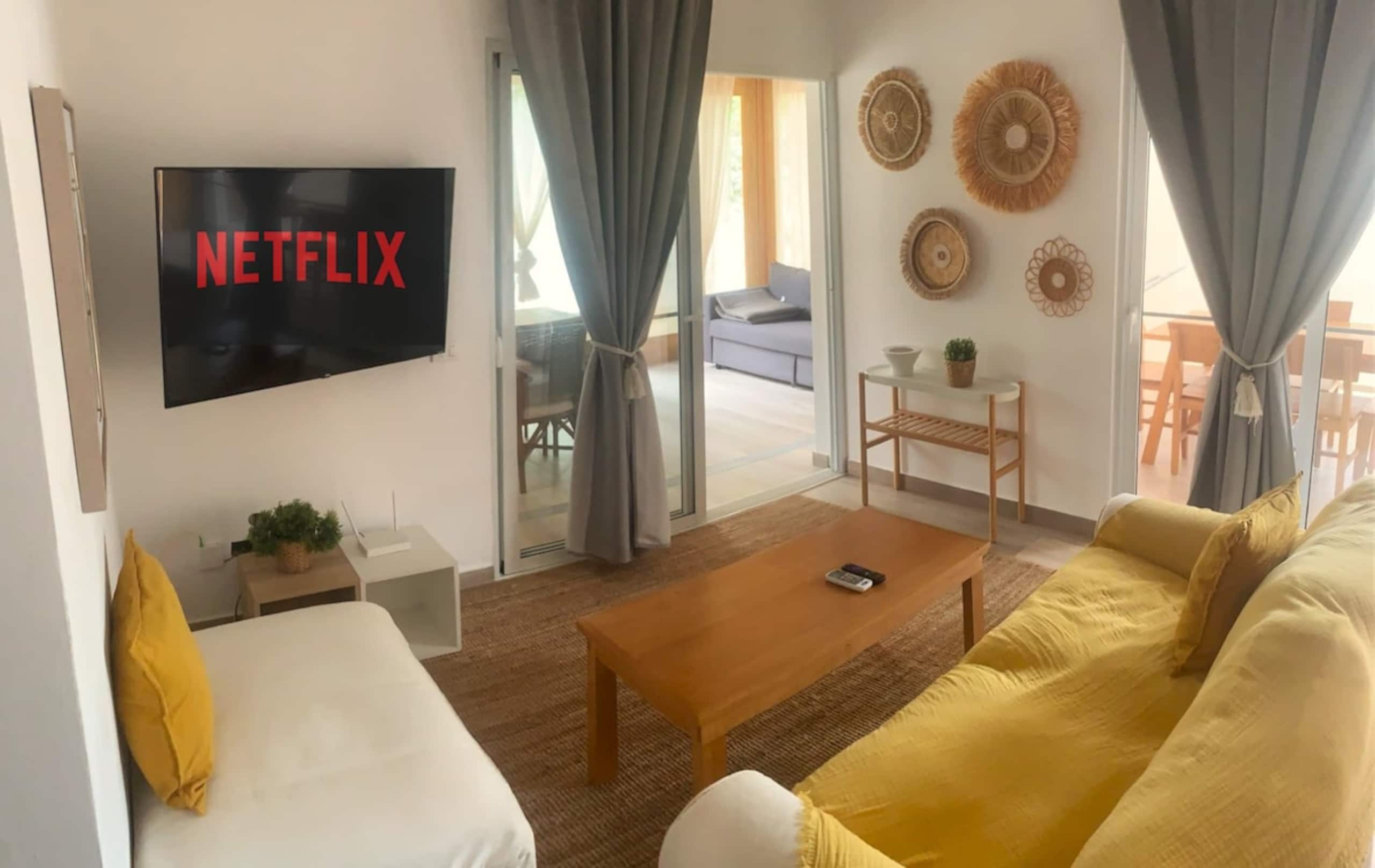 Una sala de estar con una televisión mostrando 'NETFLIX', un sofá, una mesa de centro y un sillón.