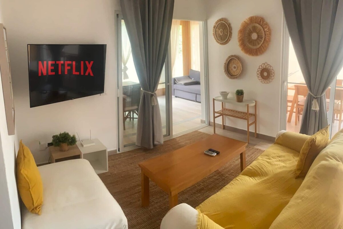 Een woonkamer met een televisie waarop 'NETFLIX' staat, een bank, een salontafel en een divan.