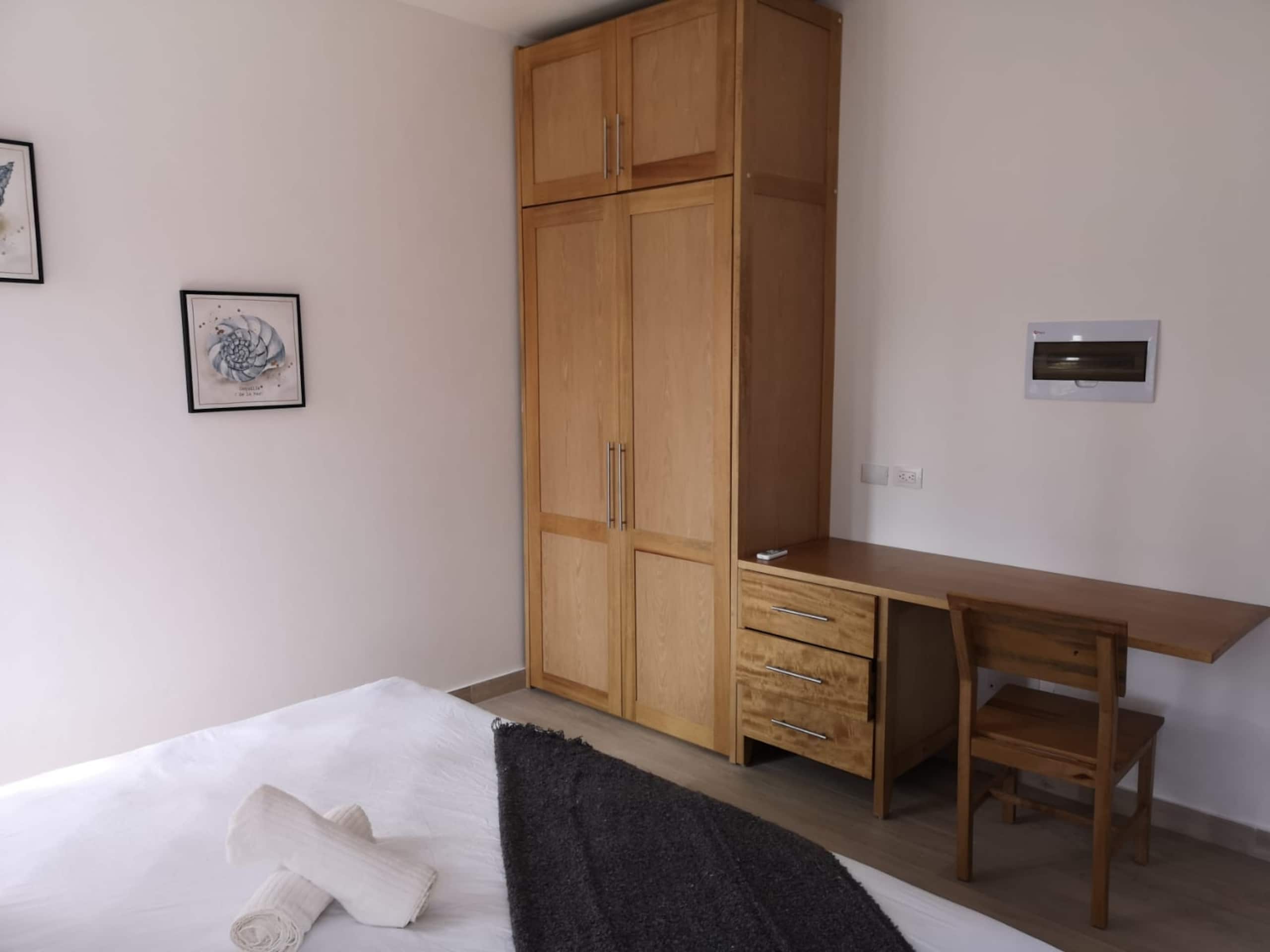 Een slaapkamer met een bed, een houten kledingkast, een bureau met een stoel en een wandairconditioner.