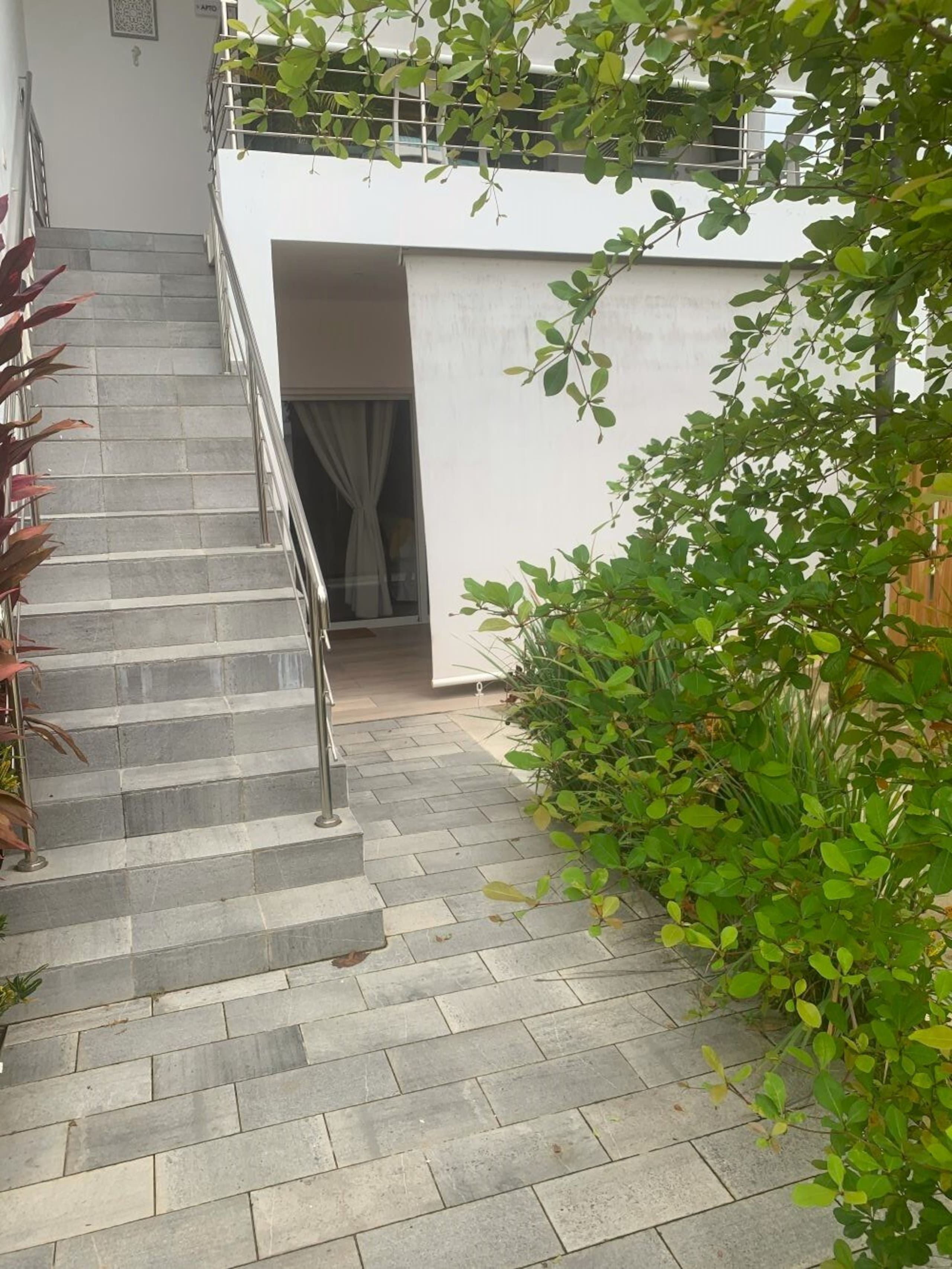 Escadaria que leva a um prédio branco com guarda-corpo de metal, cercado por vegetação e um chão de azulejos.