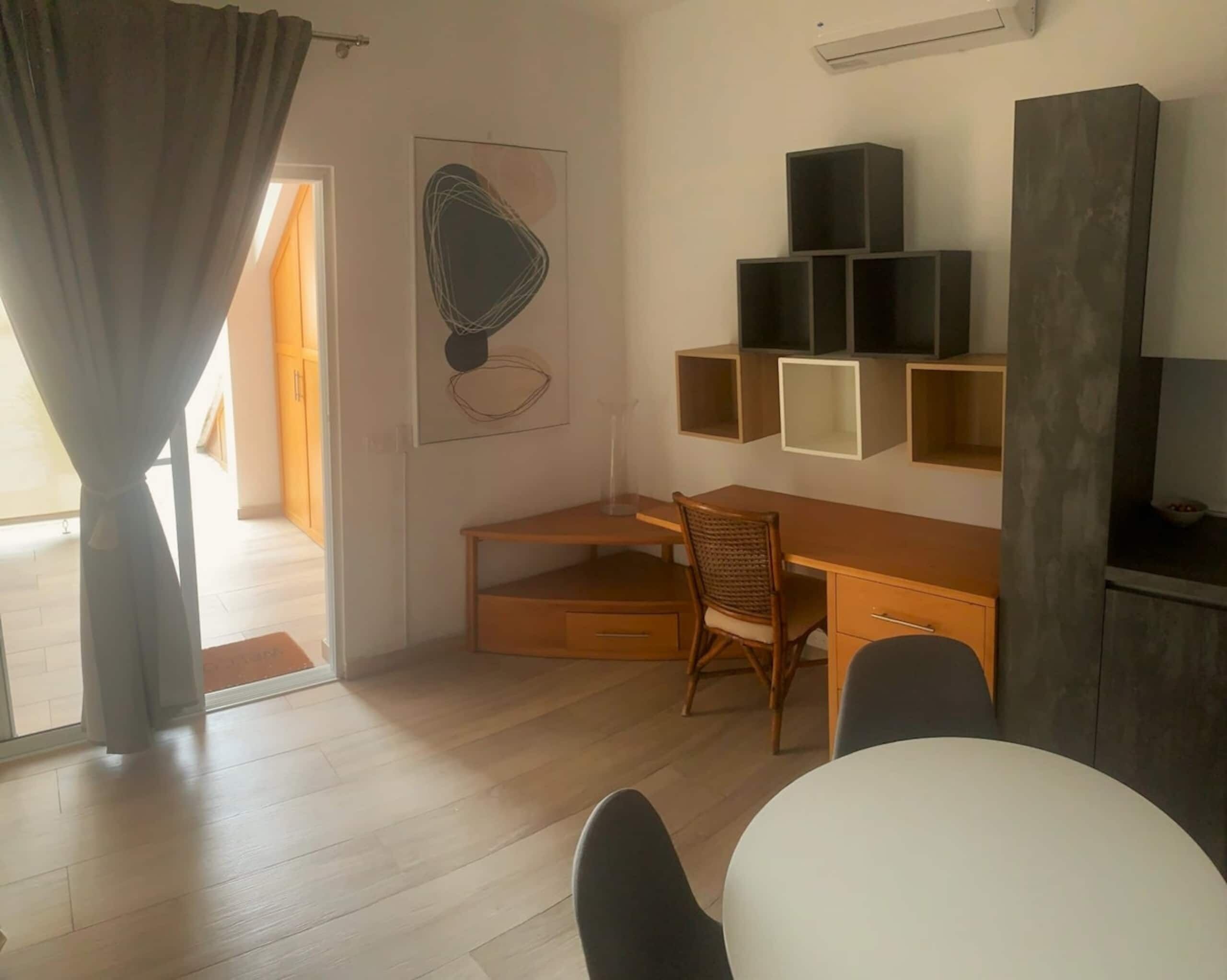 Una habitación moderna con un escritorio de madera, una silla, una mesa blanca y un ventanal con cortinas.