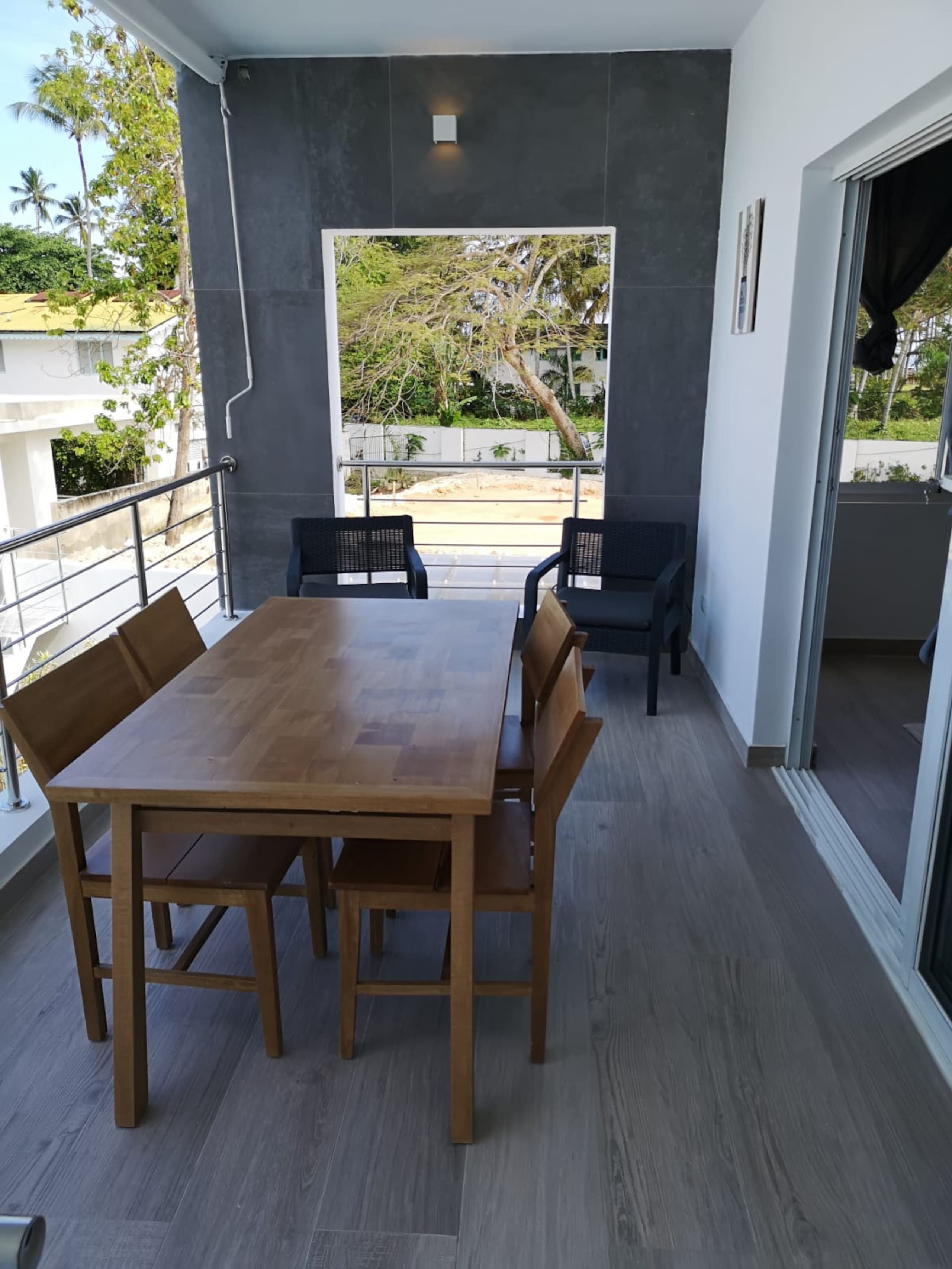 A modern dining area with a wooden table and chairs, a view of greenery, and a balcony with a railing.