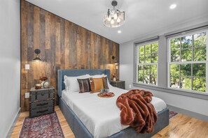 A bedroom with a large bed, wooden wall paneling, a window with a view of trees, and a chandelier. - Historic Bay Village / Cozy 1BR Getaway (Boston)