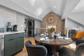 A modern kitchen with a built-in washer and dryer, a dining area with a round table and chairs, and a brick wall with a clock. - Historic Bay Village / Cozy 1BR Getaway (Boston)