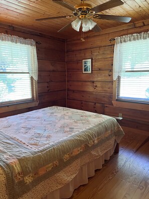 A bedroom with a wooden ceiling, a ceiling fan, a bed with a floral bedspread, and two windows with curtains. - Peaceful 3 BR + Loft river-front cabin close to Lake Lure, Chimney Rock, TIEC. (Rutherfordton)