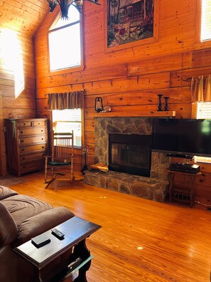 A wooden cabin interior with a stone fireplace, wooden furniture, and a television. - Peaceful 3 BR + Loft river-front cabin close to Lake Lure, Chimney Rock, TIEC. (Rutherfordton)