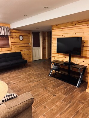 Ein Wohnzimmer mit einer schwarzen Couch, einem Fernseher auf einem Schrank, Holzwänden und Holzboden.