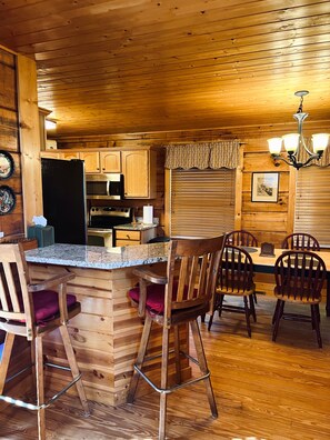 A wooden interior with a kitchen area, bar stools, and a dining table. - Peaceful 3 BR + Loft river-front cabin close to Lake Lure, Chimney Rock, TIEC. (Rutherfordton)