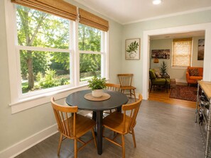 Un comedor con una mesa redonda y sillas, ventanas amplias con persianas, una planta sobre la mesa y vistas al exterior.