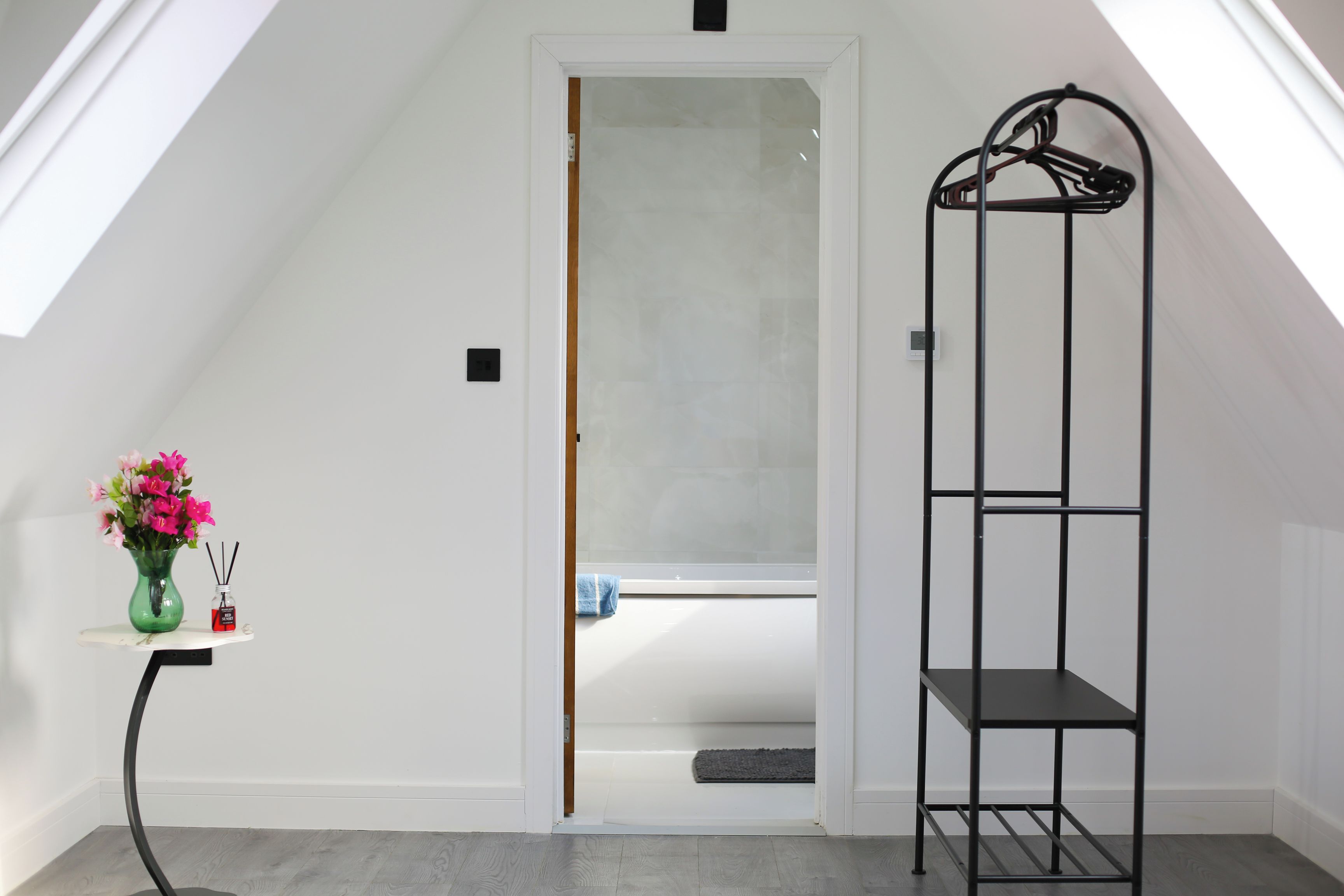 A room with a sloped ceiling, a door leading to a bathroom, a black metal shelf, and a small table with a vase of flowers.