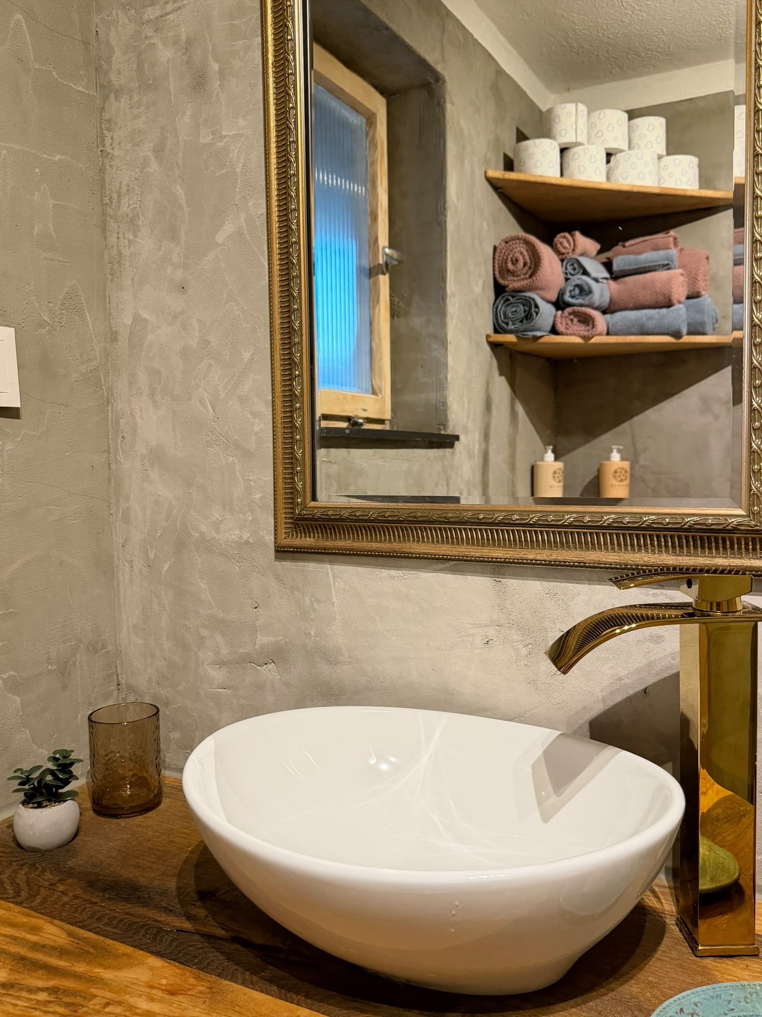 A bathroom with a white vessel sink, a gold faucet, a mirror with an ornate frame, and a shelf with towels and toiletries.