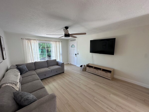 Una sala de estar con un sofá modular, un ventilador de techo, un televisor de pantalla plana y una puerta con la leyenda 'SAN CLEMENTE'.